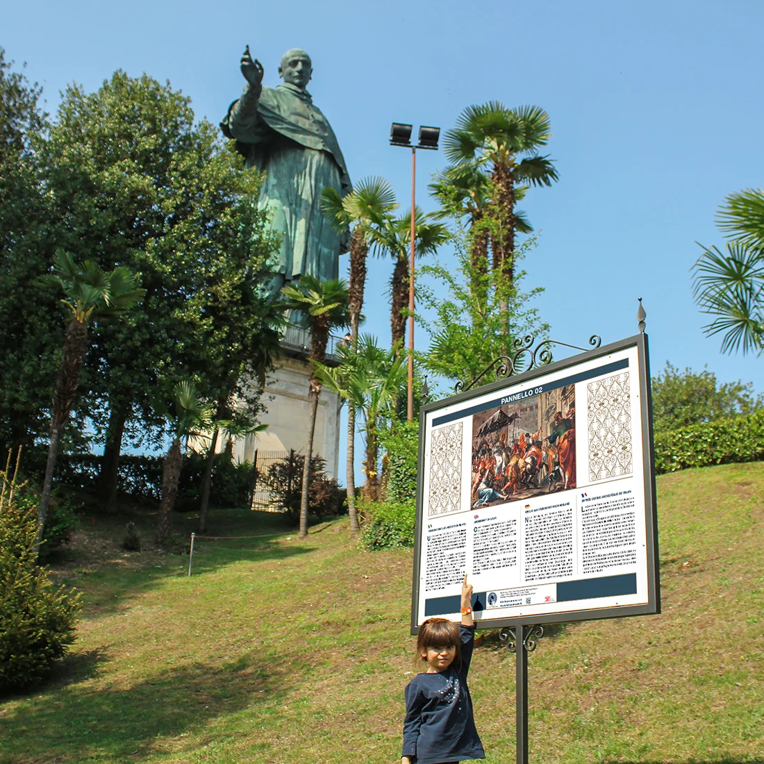 Bimba che indica uno dei pannelli informativi installati nel parco della Statua di San Carlo ad Arona, con la statua visibile sullo sfondo